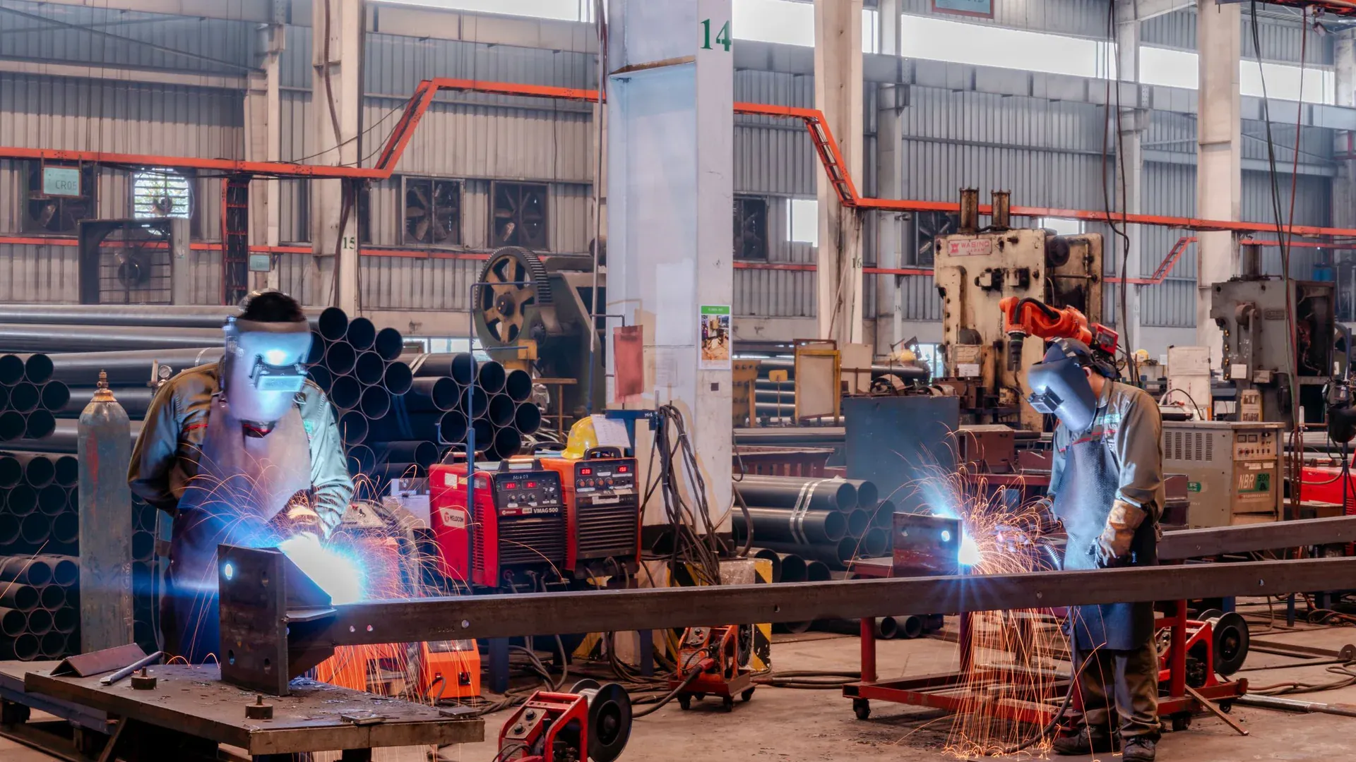 Two welders wearing protective gear working in a busy industrial factory workshop