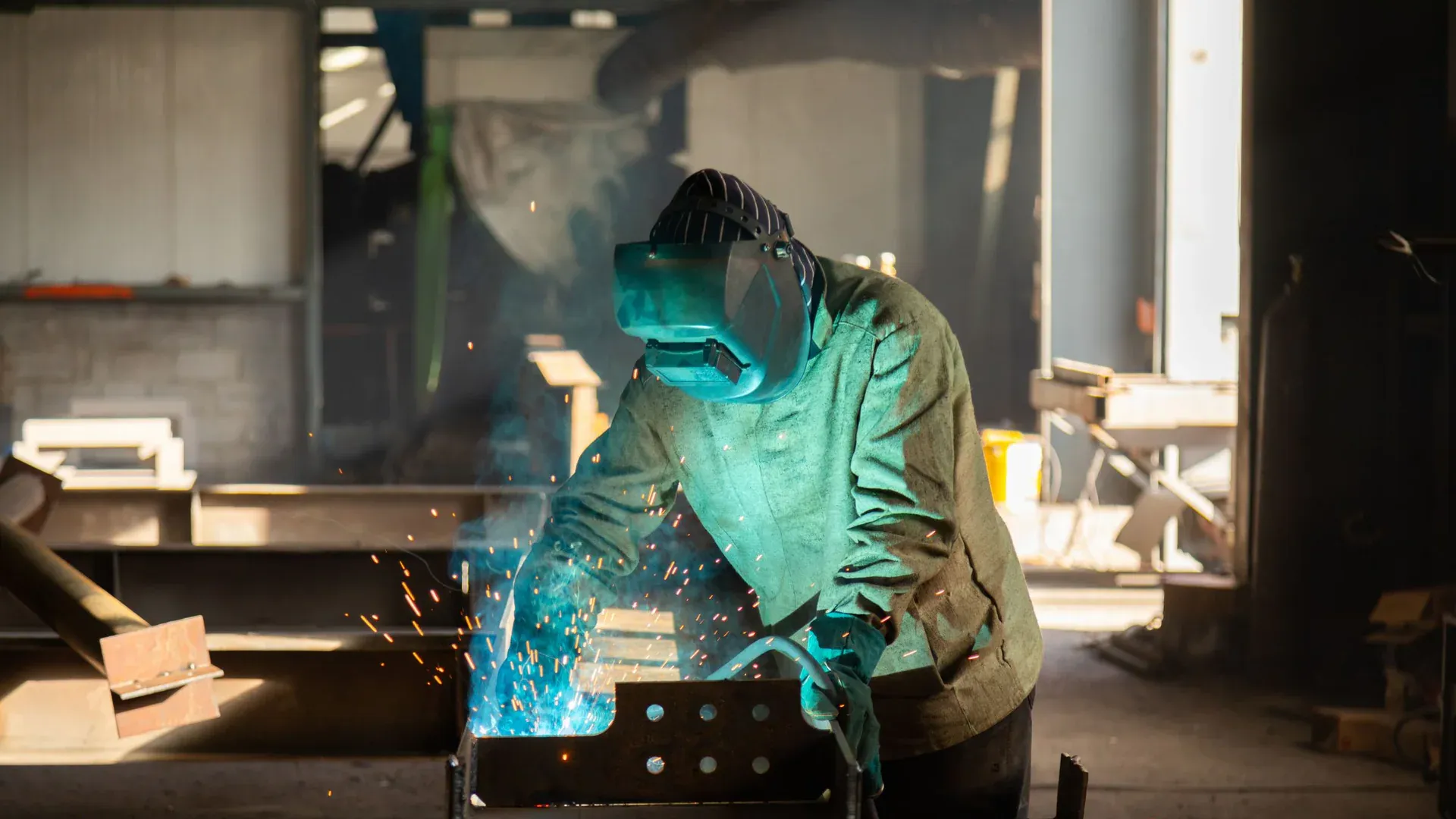 A welder wearing protective gear works in a workshop, welding metal with visible sparks
