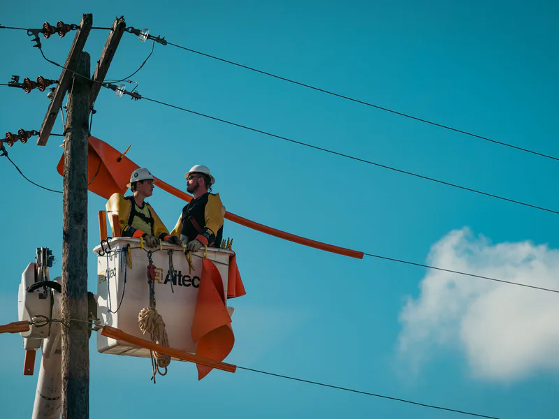 Two electricians in safety gear working on a utility pole with a clear blue sky backdrop