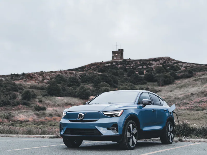 A blue Volvo electric car charging in a scenic outdoor location with a hill in the background
