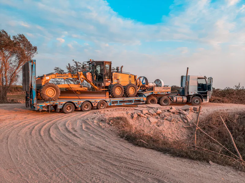 A cargo truck transporting heavy machinery on a rugged dirt road under a clear sky