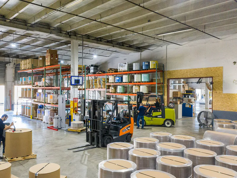 Interior view of a busy warehouse with forklifts and workers handling large rolls of material