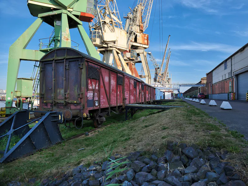 A vintage rail cargo wagon at an industrial port with cranes and warehouses